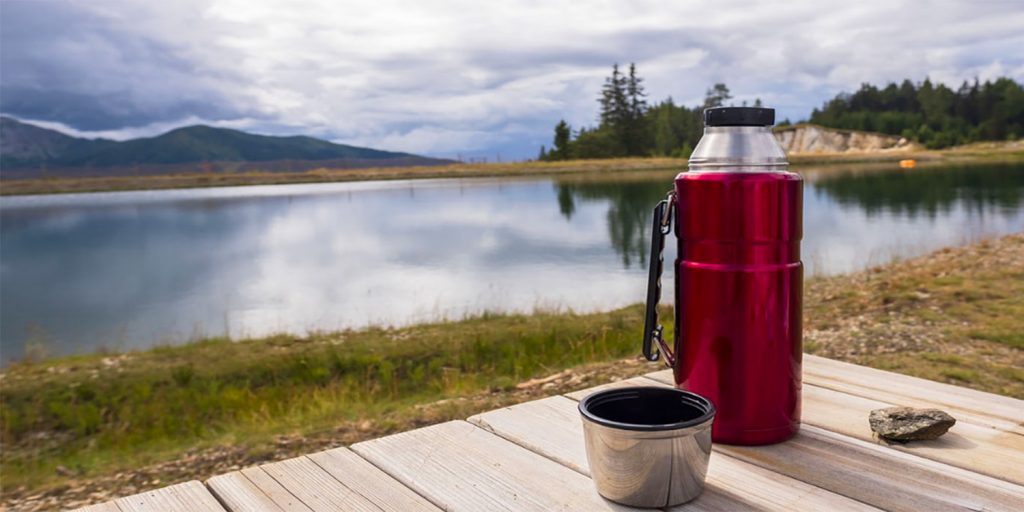 A picture of a red hydro flask on a wooden table in front of a scenic lake and grassy bank