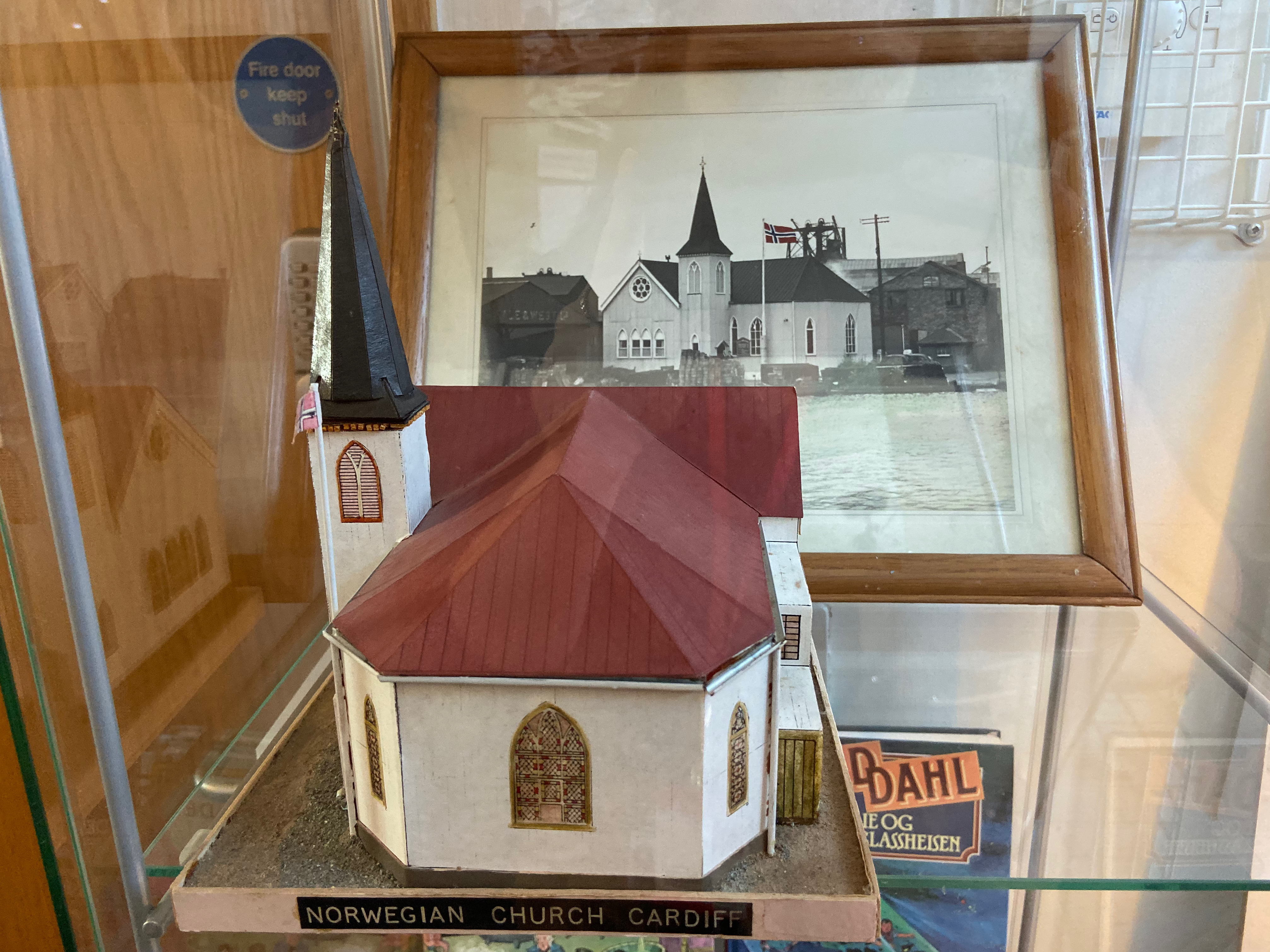A small scale model of the Norwegian Church sits in front of an old picture of the building, inside a glass case 