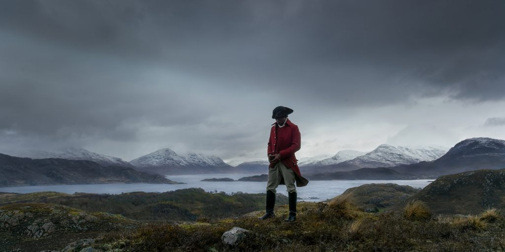 A man in historical clothing stands in front of a dramatic landscape in a work of art displayed at the National Museum Cardiff.