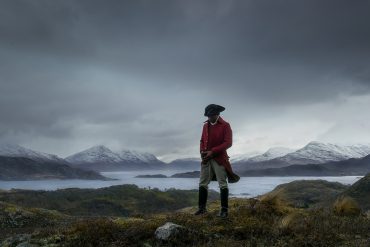 A man in historical clothing stands in front of a dramatic landscape.