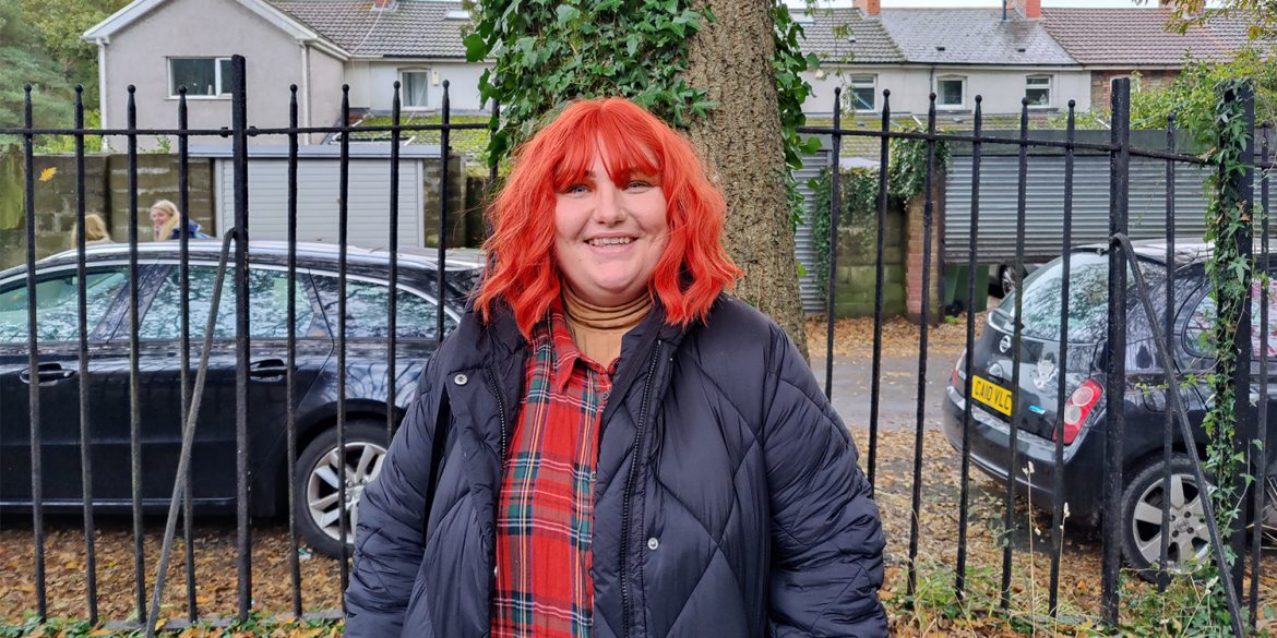Non-binary Instagrammer Dot Masters with bright red hair smiles at the camera, with trees behind them