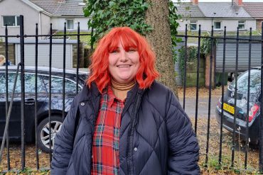 Non-binary Instagrammer Dot Masters with bright red hair smiles at the camera, with trees behind them