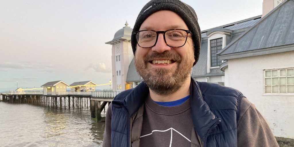 44-year-old indie cinema manager Ben Rive, a man with a brown stubbly beard, stands smiling, facing the camera, in front of Penarth Pier Pavilion.