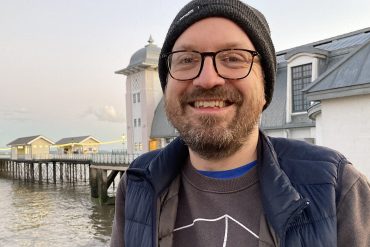 44-year-old Ben Rive, a man with a brown stubbly beard, stands smiling, facing the camera, in front of the Penarth Pier Pavilion.