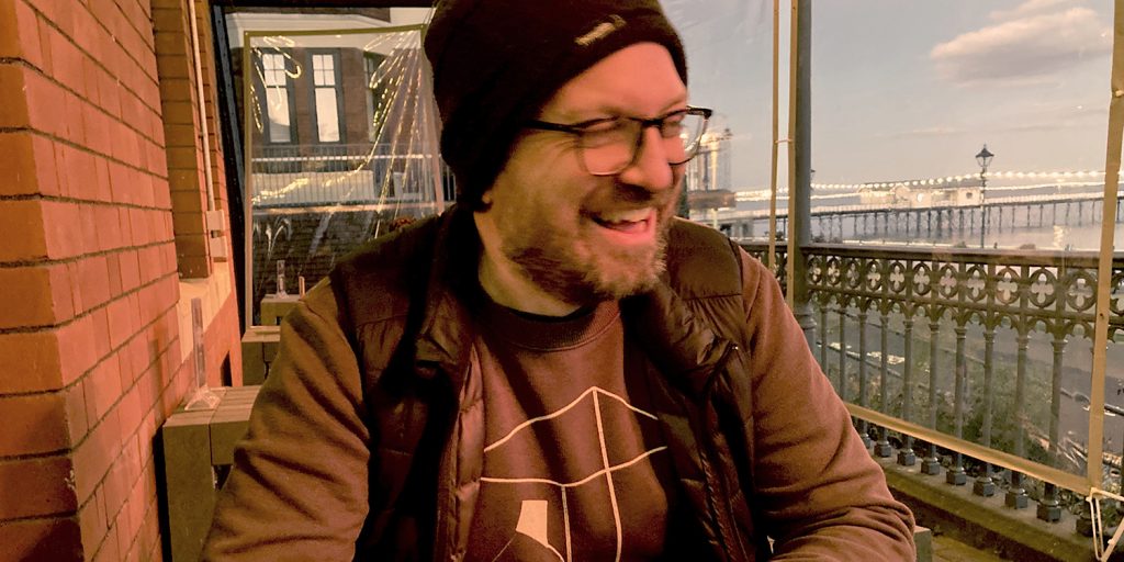 Indie cinema manager Ben Rive sits at a cafe table, laughing. He is wearing a beanie hat and brown jumper.
