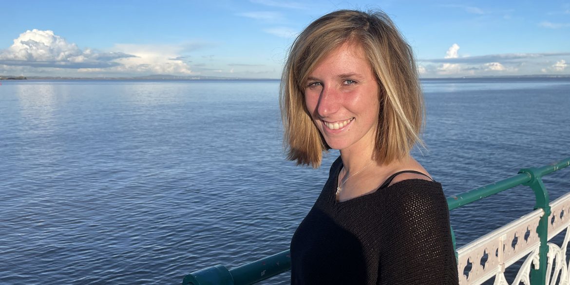 Woman in her twenties with short blonde hair smiles at the camera with the ocean behind her