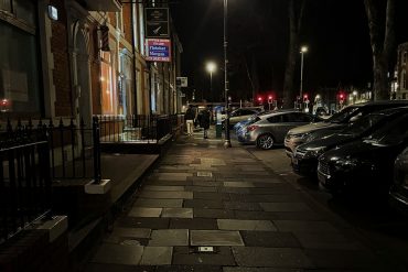 A pavement with houses on the left and cars on the right at night with light coming from street lamps