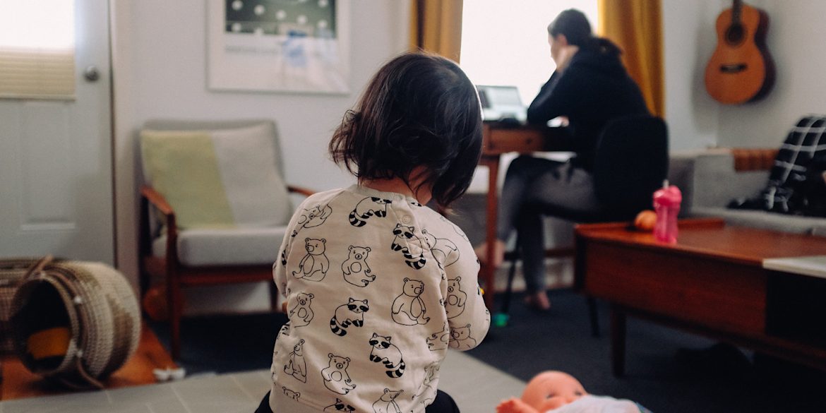 A child sits on the floor facing her mother who is sat working at a desk