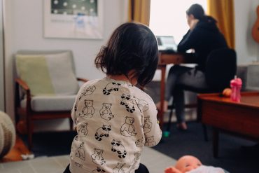A child sits on the floor facing her mother who is sat working at a desk