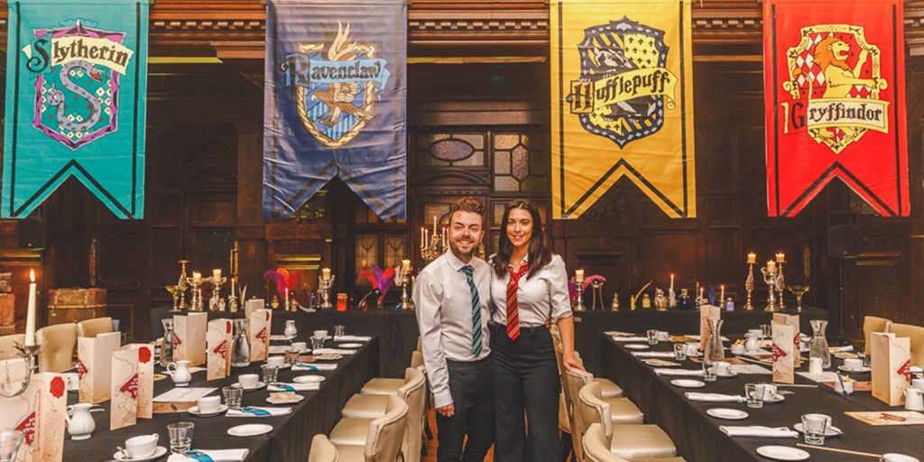 A man and a woman dressed up in Slytherin and Gryffindor uniforms, standing in front of the Hogwarts house banners in Coal Exchange Hotel for the Harry Potter afternoon