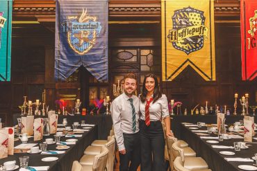 A man and a woman dressed up in Slytherin and Gryffindor uniforms, standing in front of the Hogwarts house banners in Coal Exchange Hotel
