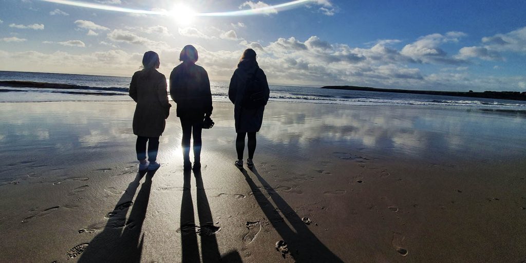 Three women standing on a beach facing away from the camera looking out to sea