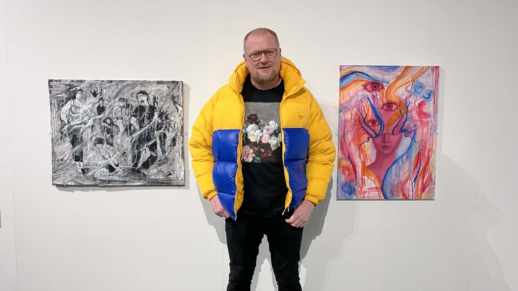 Alan Whitfield, stood centrally against a white wall in an art gallery, alongside two paintings by Tina Rogers on his left and Lia Bean to his right.