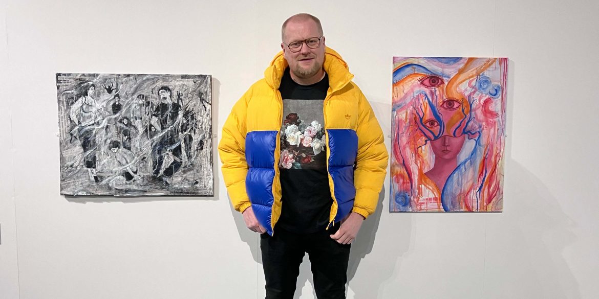 Alan Whitfield, stood centrally against a white wall in an art gallery, alongside two paintings by Tina Rogers on his left and Lia Bean to his right.