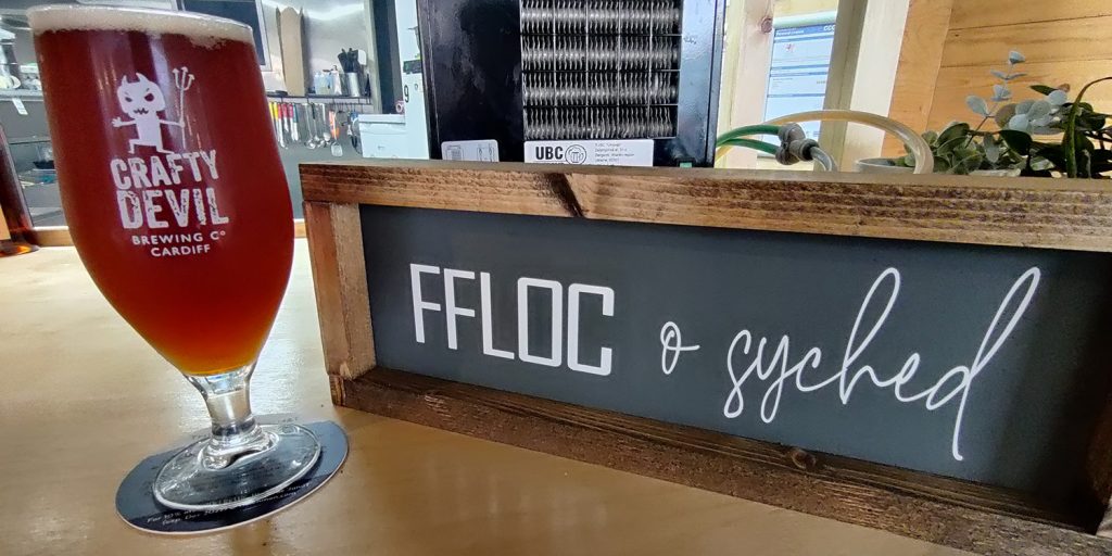 To the left: a half pint of amber ale in a teardrop glass, sat on a light wooden table. The glass has the Crafty Devil logo on it: a small, white, cartoonish devil.
To the right: a chalkboard sign that reads 'Ffloc o syched', Welsh for 'Flock of thirst'.
