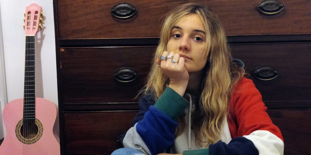 Musician Mali Hâf, dressed in a multicoloured patchwork hoodie, is sat in front of a dark wooden chest of drawers with her right hand on her chin. To the left: a pink guitar leant against a white wall.