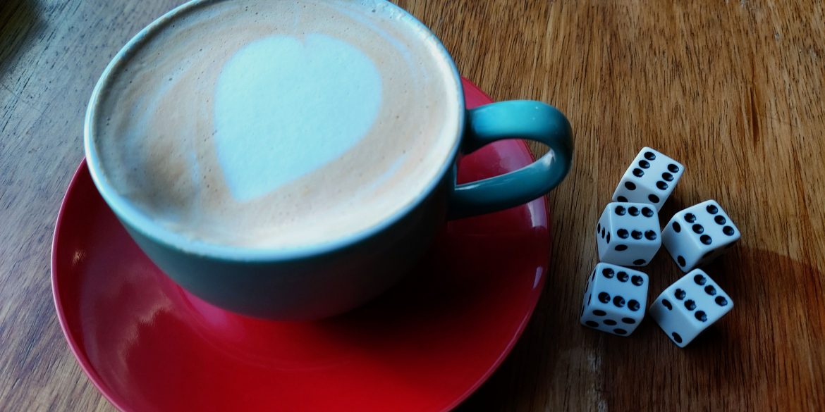 A cup of coffee in a red mug sits beside five dice in a cosy board game cafe.