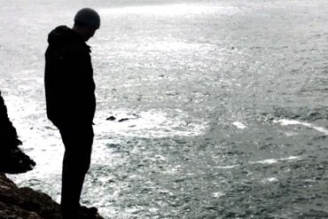 A young person stands at the edge of a cliff overlooking the sea.