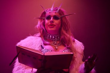The drag queen Polly Amorous is dressed in white fur and holds a large book.