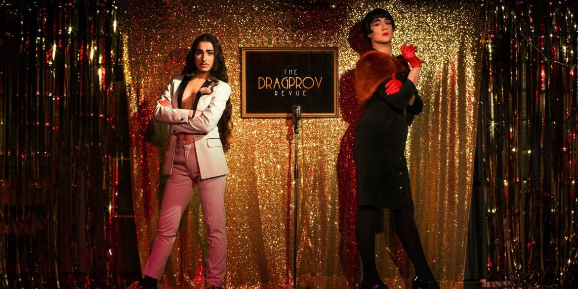 two drag artists stand on a stage with a sign between them at a drag show