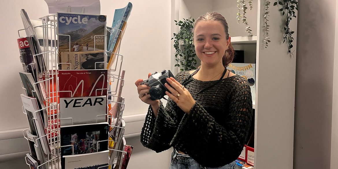 Young female photographer holds camera infant of bookshelf as she joins the creative industry in Cardiff.