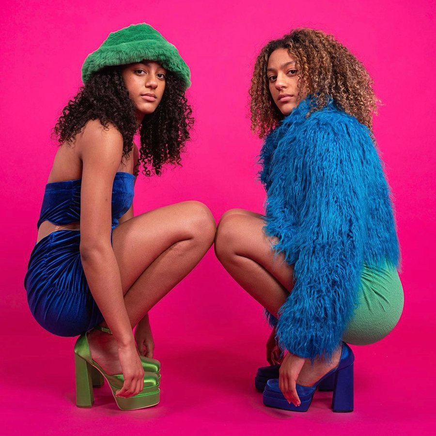 Two women crouched down dressed in green and blue against a pink backdrop representing the Cardiff creative community.