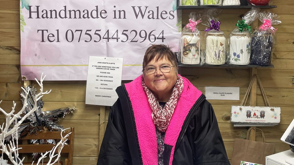 Jan sits in her Christmas stall, a sign behind her saying "Handmade in Wales"