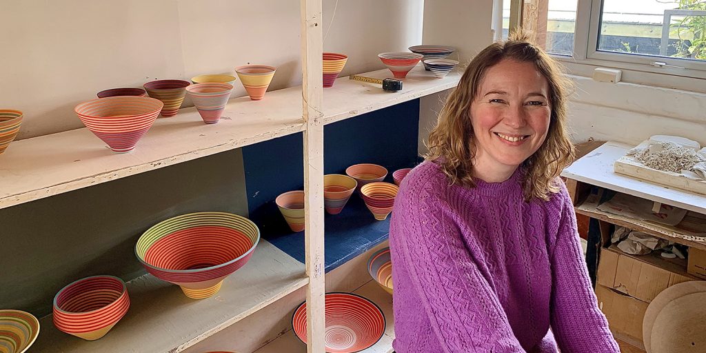Ceramicist, Sara Moorhouse sits in front of shelves of her banded ceramics 