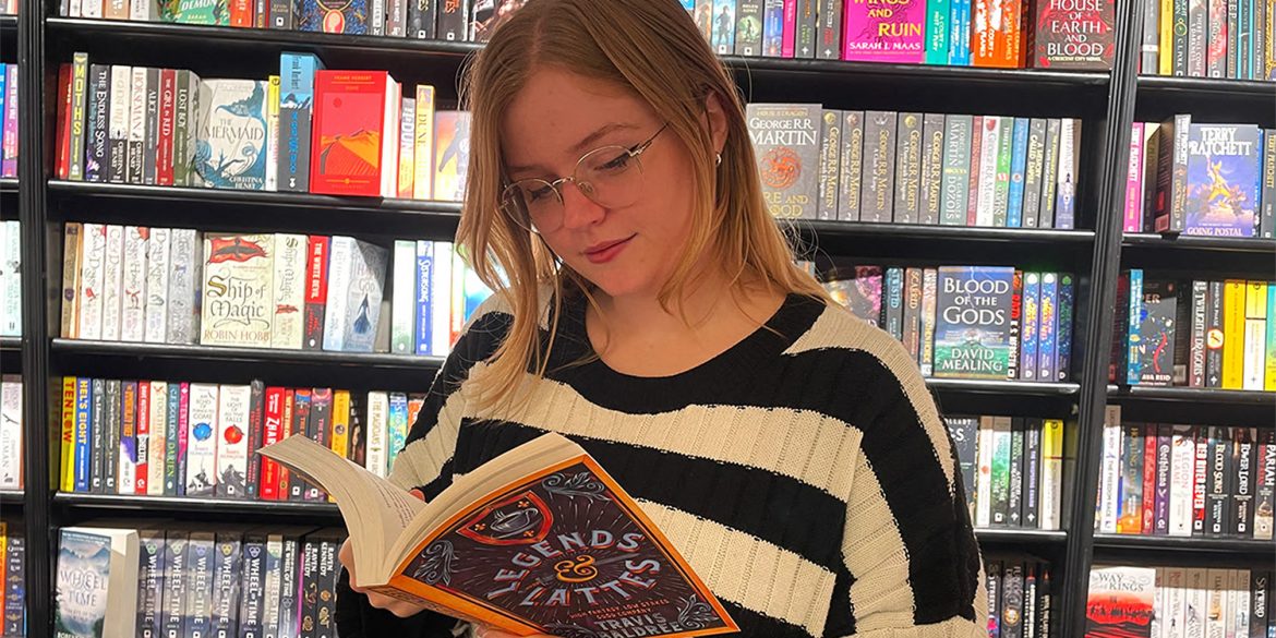 Rowan standing in front of the fantasy book section of Waterstones, reading fantasy paperback.