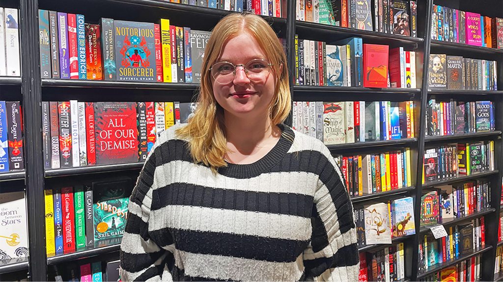 Rowan standing in front of the fantasy section of Waterstones with shelves of books behind her.