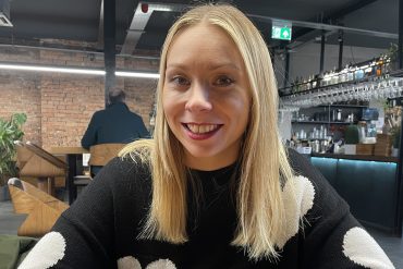 a blonde haired woman sitting at a table in a cafe, smiles at the camera.