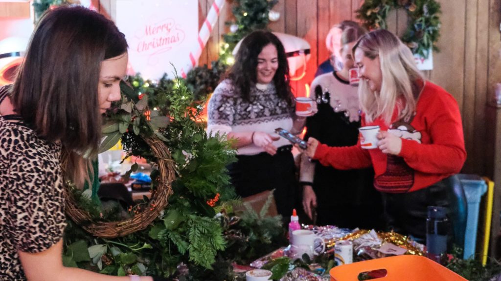 a woman holds up a wreath and looks at the items on the table while three other women stand in the background laughing and drinking mulled wine.