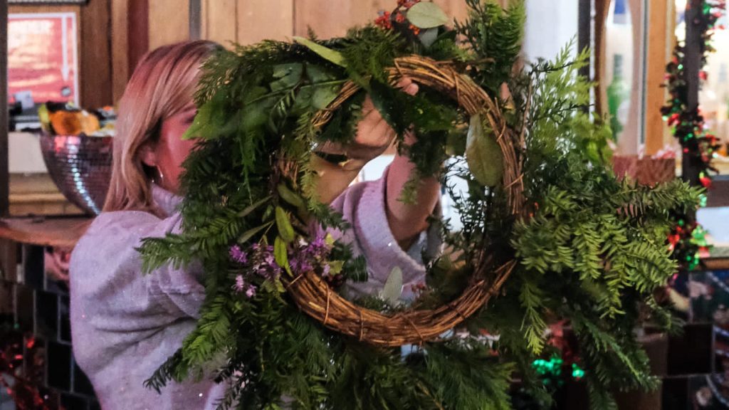 a woman with blonde hair holds a wreath up, facing her. It covers her face as she inspects it and makes adjustments.