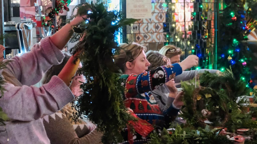 a row of women sit wearing Christmas jumpers, lifting their wreaths in the air as they work on them
