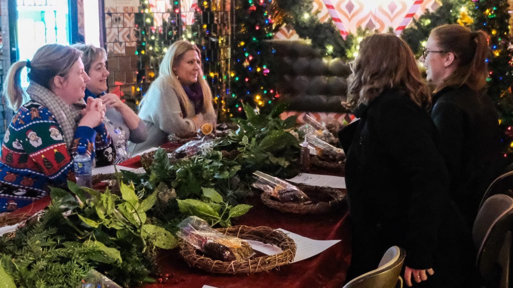 5 women sit at a table, chatting. Wreath-making materials sit in the centre of the table and bright Christmas decorations surround them.