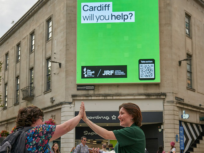A member of the public high-fives a food bank volunteer while the billboard shows a Trussell Trust campaign