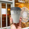 White reusable return coffee cup held by a hand in front of a cafe