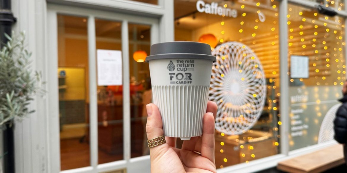 White reusable return coffee cup held by a hand in front of a cafe