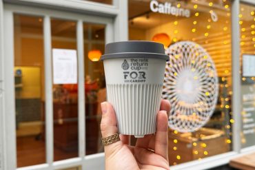 White reusable return coffee cup held by a hand in front of a cafe