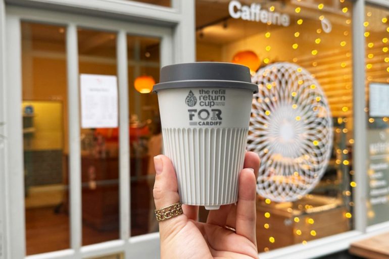 White reusable return coffee cup held by a hand in front of a cafe