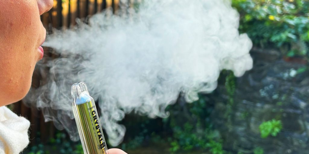 Girl using disposable vape, blowing smoke outside.