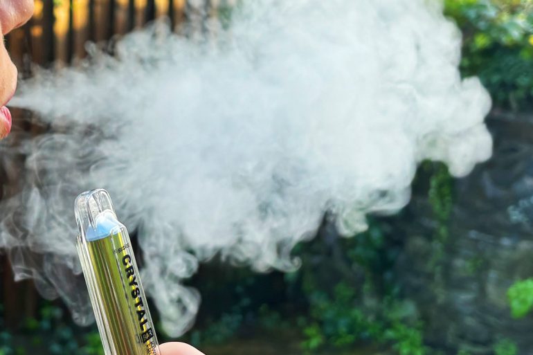 Girl using disposable vape, blowing smoke outside.