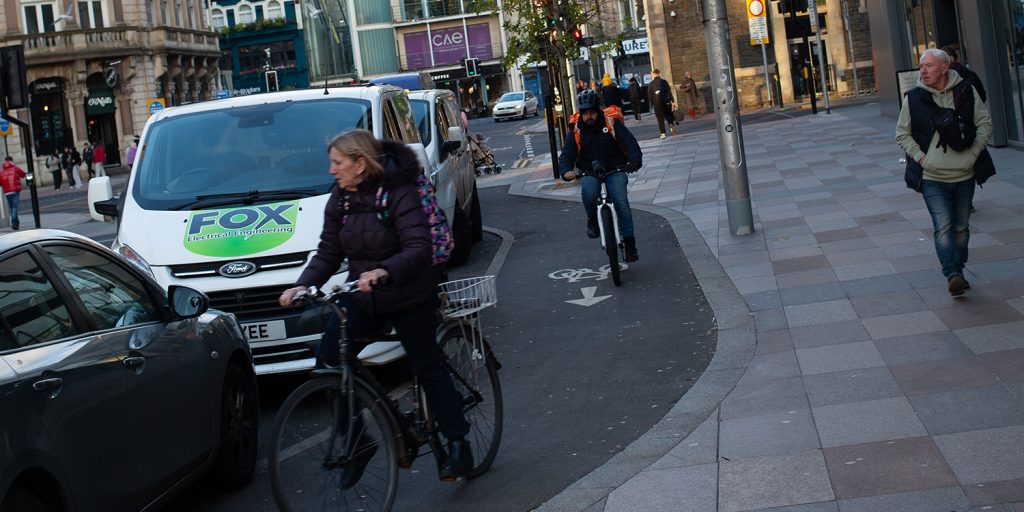 A woman is blocked by a car parked on a cycle path while a delivery cyclist comes speeding up the rear. Man looks on in wonder at the ordeal. 