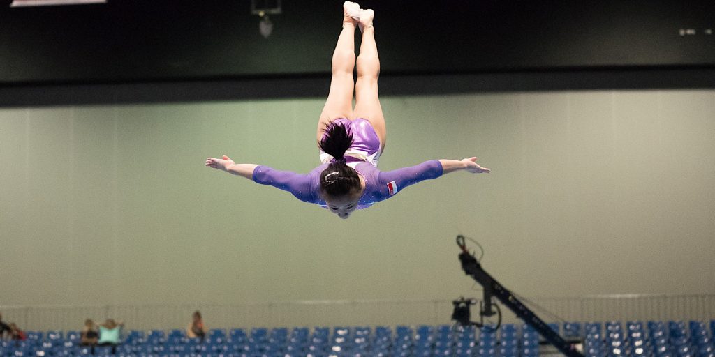 A gymnast in purple uniform is suspended in mid air as she flips on the beam
