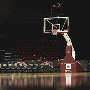 A basketball court glints in the darkness with a basketball hoop in the centre of the image