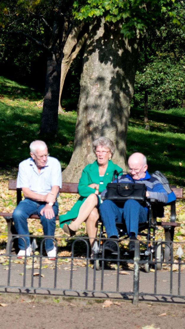 Three elderly people sitting on a park bench, a man in a white shirt on the left, a woman in a green jacket in the center, and another man in blue sitting on an electric wheelchair