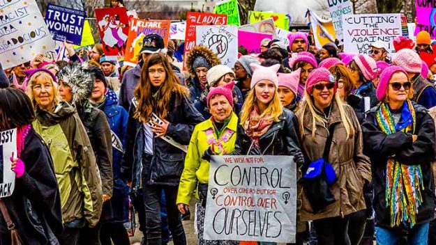 A range of women march toward the camera wearing pink hats and carrying signs