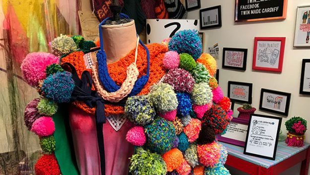 A brightly coloured pompom vest hangs over a dressmakers dummy against a background of framed embroidery projects
