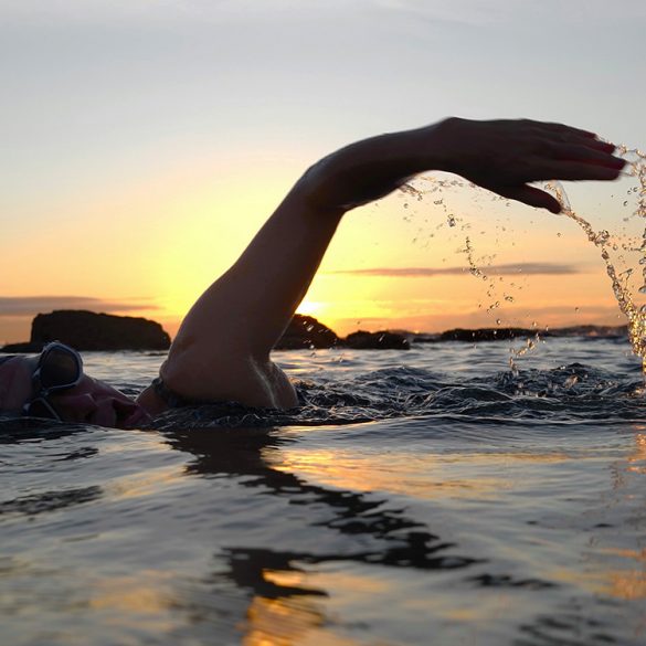 A picture of someone swimming in the sea. They are mid front crawl movement, with an arm in the air. The sun is setting behind them