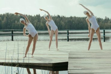Three outdoor swimmers in white swimming costumes stretch by a lake